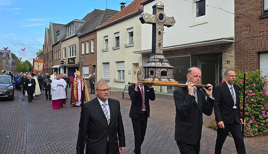 Vier Männer in schwarzen Anzügen tragen oder begleiten ein kunstvolles Kreuz durch eine Straße, im Hintergrund folgt eine Prozession. Vier Männer in schwarzen Anzügen tragen oder begleiten ein kunstvolles Kreuz durch eine Straße, im Hintergrund folgt eine Prozession.
