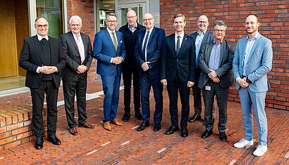 Neun Männer stehen nebeneinander vor einem Gebäude.