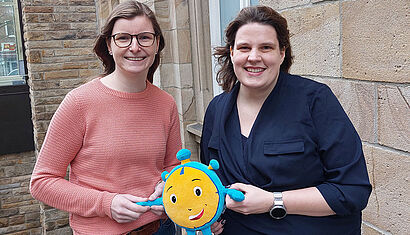 Julia Wehofsky (links) und Anna-Lena Vering halten eine Plüschfigur in der Hand, die eine Stoppuhr mit Gesicht zeigt.