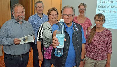 Drei Frauen und drei Männer stehen nebeneinander vor einer Leinwand, der Mann in der Mitte hält mit ausgestrecktem Arm eine Plastikflasche in der Hand.