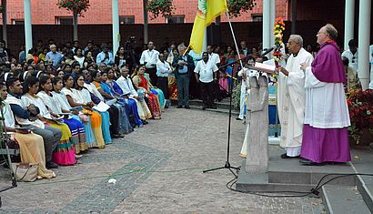 Wallfahrtsrektor Gregor Kauling (rechts) steht im Forum Pax Christi neben einem anderen Priester am Rednerpult. Vor ihm sitzen Menschen mit bunter Kleidung.