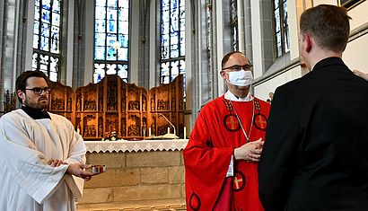 Weihbischof Rolf Lohmann steht mit Mundschutz in der Kirche, vor ihm, mit dem Rücken zum Betrachter, ein junger Mann.