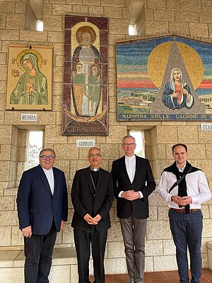 Vier Männer stehen gemeinsam vor einer Steinwand mit drei religiösen Mosaiken. Die Mosaiken zeigen jeweils Darstellungen der Mutter Gottes aus Russland, Deutschland und Italien. Die Männer tragen formelle Kleidung; drei von ihnen sind in dunklen Anzügen mit klerikalen Hemdkragen gekleidet, der vierte trägt Hemd und dunkle Hose mit einem über die Schultern gelegten Pullover. Alle lächeln in die Kamera und stehen in einem offenen, überdachten Gang aus hellem Naturstein.