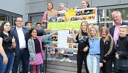 Schüler und Lehrer stehen an einer Stellwand auf der Fotos und der Schriftzug steht: "Wir haben gewonnen".