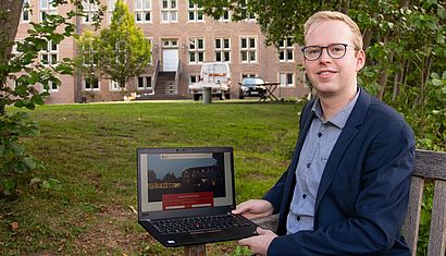 Markus Toppmöller sitzt auf einer Holzbank und hat einen Laptop auf dem Schoß, im Hintergrund ist die Fassade der Wasserburg Rindern zu erkennen.