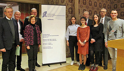 Vertreter der Jury und der Schulen stehen vor einem Banner der Gesellschaft für christlich-jüdische Zusammenarbeit Münster.