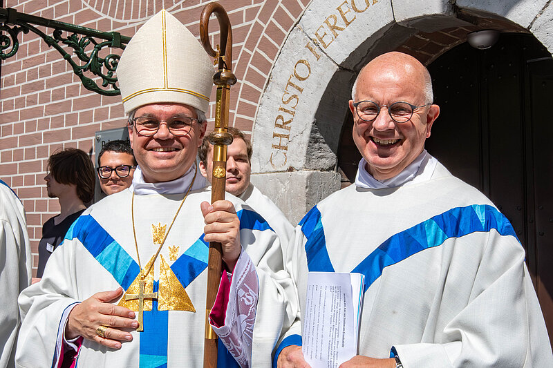 Ein Bischof und ein Priester stehen lachend vor dem Eingang des Priesterhauses
