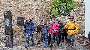 Eine Gruppe von Wandernden, darunter Dr. Heiner Wilmer SCJ,geht durch einen schmalen Durchgang zwischen alten Steinmauern. Die Menschen tragen wetterfeste Outdoor-Kleidung in verschiedenen Farben, einige nutzen Wanderstöcke. Im Hintergrund sind weitere Personen, eine historische Laterne und grüne Bäume zu sehen.