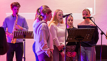Vorne rechts im Bild stehen fünf junge Frauen in einem Halbkreis um einen Notenständer herum und singen in zwei Mikrophone. Hinten links im Bild sieht man einen jungen Mann, der Saxophon spielt.