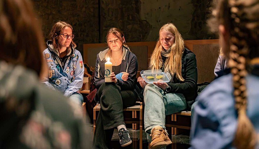 Drei junge Frauen sitzen in einem überwiegend dunklen Raum und blicken auf eine brennende Kerze.