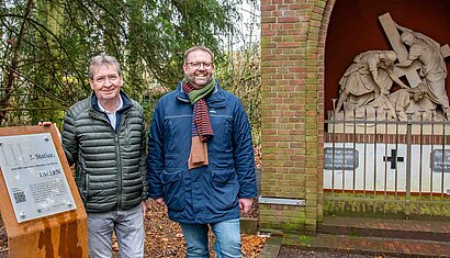 Zwei Männer stehen zwischen einer Metallstele und einer Kreuzwegstation