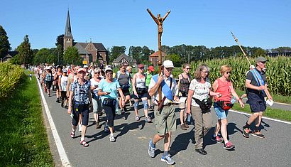 Unter strahlend blauem Himmel läuft eine große Menge Pilger über eine Landstraße, im Hintergrund ist eine Kirche zu sehen.