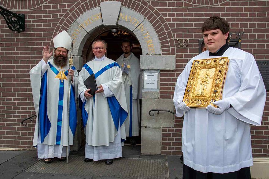 Ein Bischof und ein Priester stehen vor einem Haus, rechts steht ein Messdiener, der ein golden eingebundenes Buch trägt.