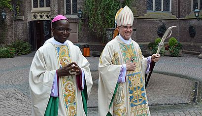 Erzbischof Lompo und Weihbischof Lohmann gehen in liturgischer Kleidung über den Brunnenhof in Kevelaer.