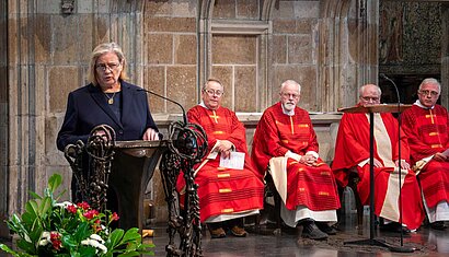 Eine Frau steht am Ambo einer Kirche, seitlich hinter ihr sitzen mehrere Priester und hören ihr zu.