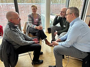 Vier erwachsene Personen sitzen in einem Stuhlkreis in einem hellen Raum mit großen Fenstern. Sie unterhalten sich miteinander, wirken aufmerksam und zugewandt. Eine Person spricht mit Gesten, während die anderen zuhören und teilweise lächeln. Im Hintergrund sind eine Backsteinmauer und Bäume zu sehen.