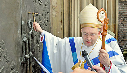 Bischof Helmut Dieser schlägt mit einem goldenen Hammer, den er in der rechten Hand hält gegen das Pilgerportal in Kevelaer. In der linken Hand hält der Bischof seinen Bischofsstab.