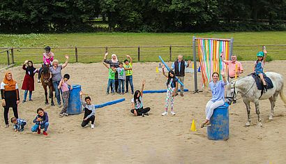 Kinder und Erwachsene stehen auf einer Reitanlage, zwei Kinder sitzen auf einem Pferd.