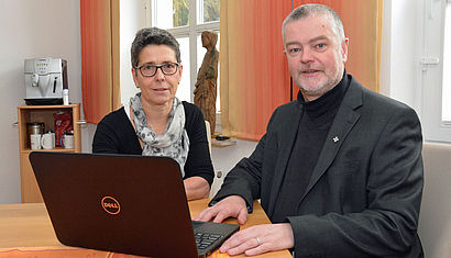 Sigrun Bogers und Dechant Stefan Keller sitzen im Pfarrbüro an einem aufgeklappten Laptop.