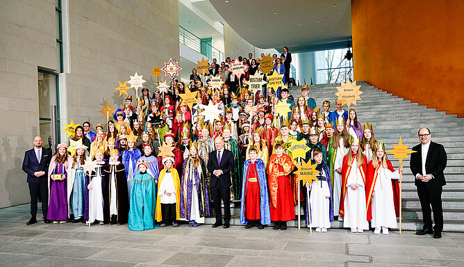 Das Bild zeigt den Empfang der Sternsinger im Bundeskanzleramt 2025. Die Kinder stehen auf einer Treppe und halten Sterne auf denen der Name des jeweiligen Bistums geschrieben steht. In der Mitte steht der ehemalige Bundeskanzler Olaf Scholz.