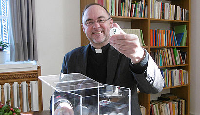 Weihbischof Rolf Lohmann sitzt vor einer Lostrommel und hält in der linken Hand einen weißen Ball, der mit der Nummer 7 beschriftet ist.