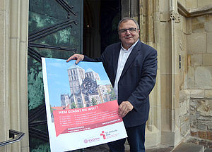 Hans-Bernd Köppen steht vor der Eingangtür zum St.-Paulus-Dom. In den Händen hält er ein Plakat, das die DomGedanken 2022 ankündigt.