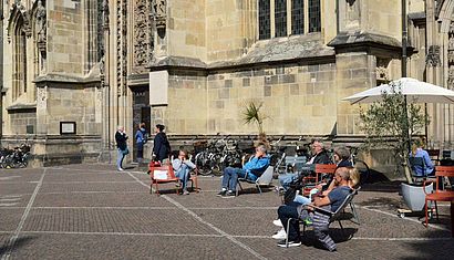 Auf dem Platz vor der Lambertikirche sitzen mehrere Menschen auf Stühlen in der Sonne.