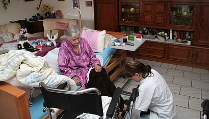 Eine Pflegerin kniet vor einer Seniorin, die in einem Wohnzimmer auf einem Pflegebett sitzt.