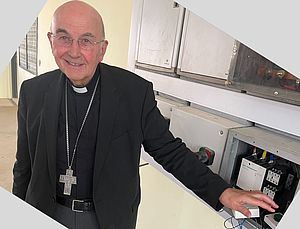 Bischof Felix Genn steht neben einem geöffneten Elektro-Schaltkasten. Seine Hand liegt auf einem Schalter.