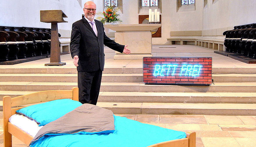 Der Superintendent wirbt mit einem Bett in der Apostelkirche für die Privatquartierkampagne.