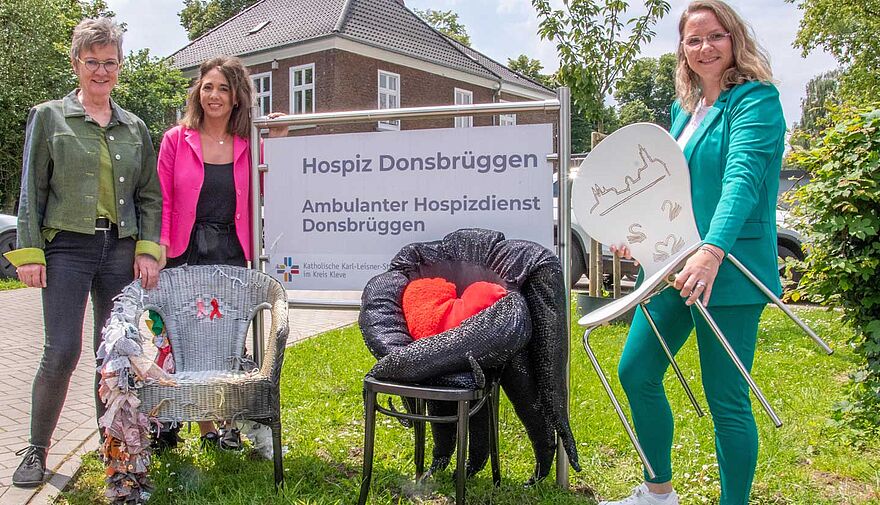 Drei Frauen stehen mit kunstvoll verzierten Stühlen vor einem Schild, auf dem u.a. "Ambulanter Hospizdienst Donsbrüggen" steht.