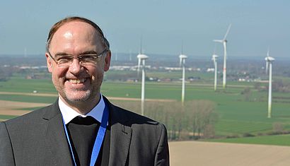 Weihbischof Rolf Lohmann steht in der Sonne, im Hintergrund ist eine niederrheinische Landschaft mit Windrädern zu erkennen.