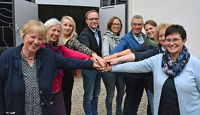 Neun Menschen stehen im Halbkreis und lachen den Betrachter an, sie strecken jeweils einen Arm in die Mitte und haben die Hände übereinander gelegt.