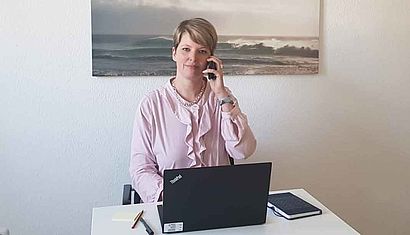 Eine Frau sitzt am Computer und hält sich mit der linken Hand einen Telefonhörer ans Ohr.