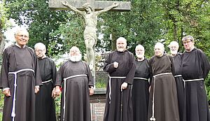 Acht mit der Tracht des Kapuzinerordens gekleidete Männer stehen vor einem steinernem Kreuz mit Christus-Corpus in einem Garten.