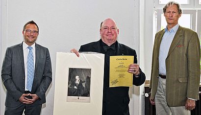 Bürgermeister Thomas Goerts, Dr. Udo Grote, der einen Kunstdruck und eine Gutschein hält, und Wilhelm Albert Herzog von Urach stehen nebeneinander.