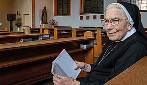 Eine ältere Ordensschwester sitzt in einer Kirchenbank und hält ein kleines Notizbuch in den Händen. Sie trägt eine schwarze Tracht mit weißem Schleier und eine Brille. Im Hintergrund sind weitere Kirchenbänke, ein Altarbereich und eine Statue zu sehen.