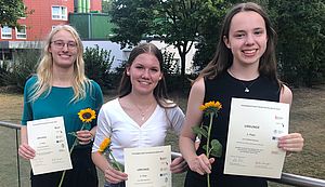 Drei junge Frauen stehen nebeneinander. Jede hält eine Urkunde und eine Sonnenblume in den Händen.