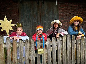 Vier Mädchen stehen hinter einem alten Holzzaun vor einer dunklen Scheunentür. Sie tragen bunte Kostüme und Kronen, wie die Heiligen Drei Könige. Das Mädchen links im Bild trägt einen großen gelben Stern. Alle lächeln freundlich. Vier Mädchen stehen hinter einem alten Holzzaun vor einer dunklen Scheunentür. Sie tragen bunte Kostüme und Kronen, wie die Heiligen Drei Könige. Das Mädchen links im Bild trägt einen großen gelben Stern. Alle lächeln freundlich.