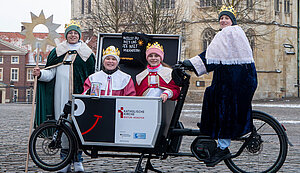 Vier als Sternsinger verkleidete Kinder stehen auf einem Lastenfahrrad vor dem St.-Paulus-Dom. Sie tragen Kronen und farbige Gewänder in Grün, Rosa und Blau. Ein Kind hält einen großen Stern auf einer Stange. Auf einem Schild am Fahrrad steht „Willst du mit uns die Welt verändern?“ sowie das Logo des katholischen Bistums Münster. Vier als Sternsinger verkleidete Kinder stehen auf einem Lastenfahrrad vor dem St.-Paulus-Dom. Sie tragen Kronen und farbige Gewänder in Grün, Rosa und Blau. Ein Kind hält einen großen Stern auf einer Stange. Auf einem Schild am Fahrrad steht „Willst du mit uns die Welt verändern?“ sowie das Logo des katholischen Bistums Münster.