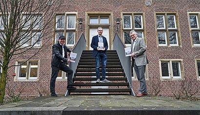 Drei Männer stehen an der Treppe der Wasserburg und halten je ein Buch in der Hand.