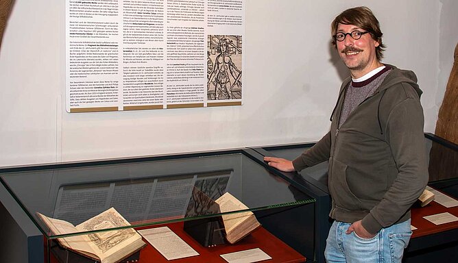 Ein Mann steht an einer Glasvitrine in der alte Bücher liegen.
