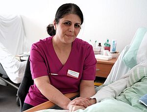 Eine mit einer Pflege-Uniform bkleidete Frau sitzt an einem Krankenbett und hält die Hand eines in dem Bett liegenden Patienten.