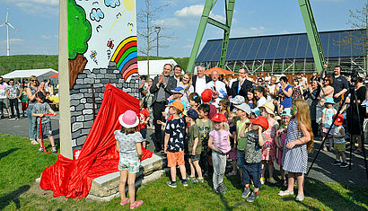 Links im Bild steht ein Stück der Berliner Mauer, im unteren Teil noch verhüllt durch ein rotes Tuch. Davor stehen Kinder. Auf der der Mauer ist ein Bild einer gemalten Mauer zu erkennen, hinter der ein Regenbogen zu sehen ist. 