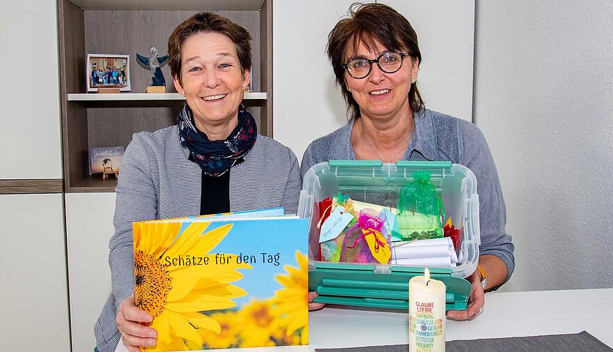 Zwei Frauen sitzen an einem Tisch, eine hält ein Buch, die andere eine Plastikbox mit bunten Inhalt.