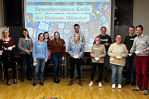 Eine Gruppe junger Menschen steht mit lächelnden Gesichtern vor einer Leinwand, auf die die Worte "Bewerber:innen-Kreis des Bistums Münster" projiziert sind.