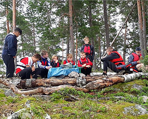 Mehrere Jungen und ein Betreuer sitzen in einem Wald auf Baumstämmen.