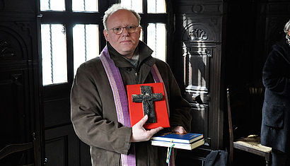 Wallfahrtsrektor Gregor Kauling steht in der Gnadenkapelle in Kevelaer, in der rechten Hand hält er ein auf einer roten Platte befestigtes, dunkles Holzkreuz.