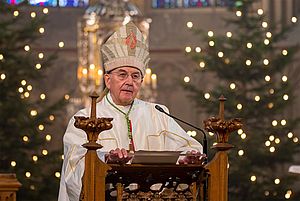 Bischof Dr. Felix Genn steht am Ambo der mit Tannenbäumen und Kerzen weihnachtlich geschmückten St-Lamberti-Kirche.