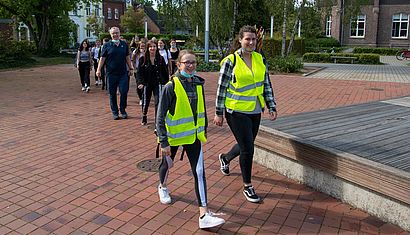 Zwei Schülerinnen in gelben Warnwesten laufen vorbei, gefolgt von einem Mann und weiteren Schülerinnen.